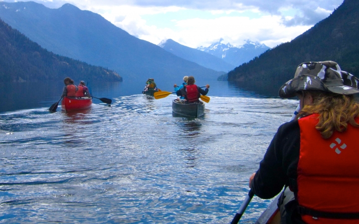 a group of outward bound students paddle canoes on calm water surrounded blue mountains